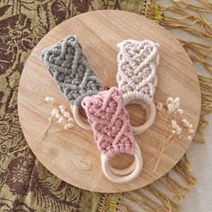 Può includere: Tre anelli per dentizione per bambini in macramè grigio, rosa e crema, ciascuno con un anello di legno. Gli anelli sono disposti su una tavola rotonda di legno, con fiori secchi e un tessuto con frange sullo sfondo.
