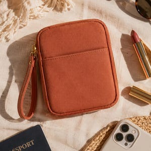 May include: A rectangular, rust-colored zippered pouch with a matching wrist strap. The pouch features a smooth, solid-colored exterior with a single front pocket and a gold-toned zipper closure, designed for carrying small personal items.
