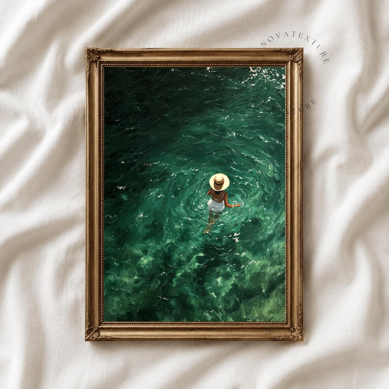 May include: A framed wall art print featuring an aerial view of a person wearing a straw hat and white swimwear floating in deep emerald green water. The frame is metallic gold with a decorative border, displayed against a white fabric background.