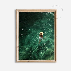 May include: A framed wall art print featuring an aerial view of a person wearing a white swimsuit and a tan straw hat, floating in deep emerald green water with visible ripples and light reflections on the surface of the clear ocean water.
