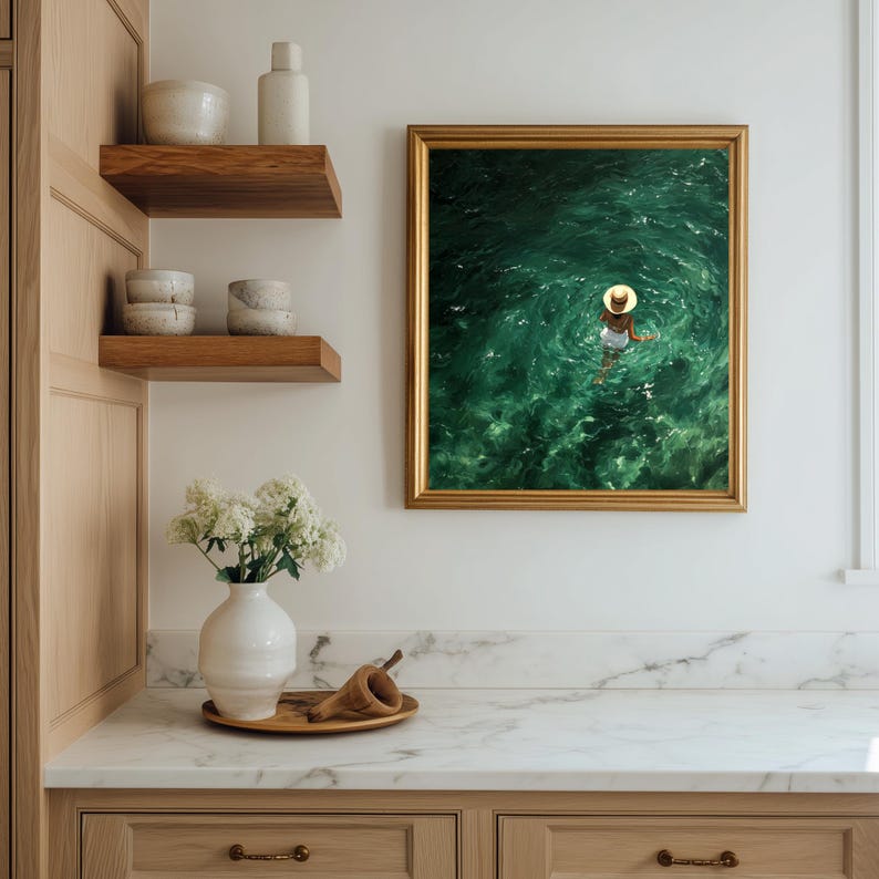 May include: A framed wall art print featuring an aerial view of a person wearing a straw hat and white swimwear floating in deep green ocean water. The artwork is displayed in a gold frame on a white wall next to wooden shelves with ceramic bowls.