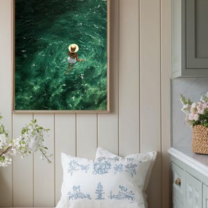 May include: A framed wall art print depicting a person wearing a straw hat and white clothing swimming in deep green water. Below, a white throw pillow features a blue line-art pattern of various figures and objects on a wooden bench.