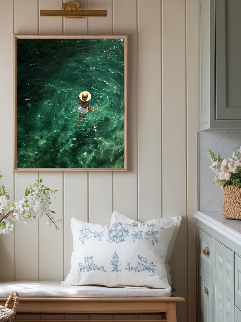 May include: A framed wall art print depicting a person wearing a straw hat and white clothing swimming in deep green water. Below, a white throw pillow features a blue line-art pattern of various figures and objects on a wooden bench.