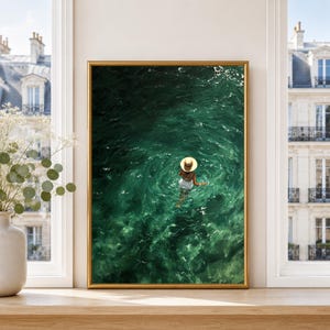 May include: A framed wall art print depicting a person wearing a straw hat and white clothing, partially submerged in deep green, rippling water. The artwork is displayed in a gold-coloured frame, positioned on a wooden surface near a window.