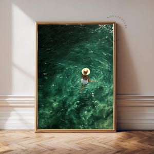 May include: A framed wall art print featuring a high-angle view of a person wearing a white swimsuit and a straw hat, floating in deep emerald green water. The water surface shows ripples and light reflections, with the text NOVATEXTURE visible.