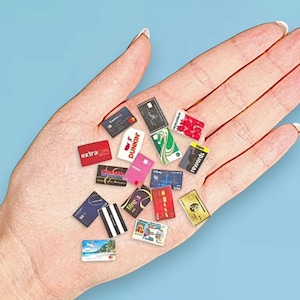 May include: A collection of miniature plastic cards featuring various brand logos and designs, including red, black, white, blue, green, and gold colors. The assortment includes small replicas of credit cards, loyalty cards, and identification cards.