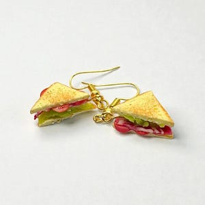 Pu&ograve; includere: Un paio di orecchini a forma di triangoli di sandwich in miniatura. Gli orecchini presentano ganci dorati e repliche dettagliate di sandwich con pane tostato, lattuga, pomodoro e quello che sembra prosciutto o pancetta.