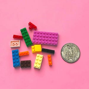 Könnte beinhalten: Eine Sammlung verschiedener Kunststoff-Bausteine in verschiedenen Farben wie Pink, Grün, Blau, Gelb, Orange, Schwarz und Rot, platziert neben einer US-Quarter-Münze als Größenvergleich, die einen Durchmesser von etwa 2,4 cm aufweist.