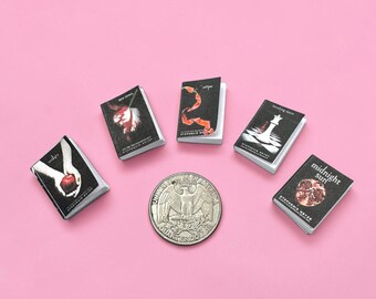 May include: Five miniature books by Stephenie Meyer arranged in a semi-circle around a United States quarter coin. The books feature black covers with distinct graphic illustrations and titles including Twilight, New Moon, Eclipse, Breaking Dawn, and Midnight Sun.