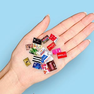 May include: A collection of miniature credit cards and store loyalty cards in various colors, including red, pink, and blue. The cards feature logos from popular retailers and brands. The cards are held in an open hand against a light blue background.