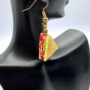 Pu&ograve; includere: Un singolo orecchino color oro con un ciondolo a forma di sandwich in miniatura. Il sandwich ha una forma triangolare con strati rossi, verdi e gialli. L'orecchino &egrave; esposto su una testa di manichino nera.