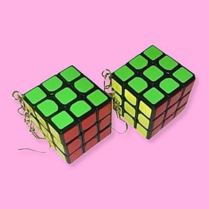 May include: A pair of Rubik's Cube earrings. Each cube has a classic colour scheme of green, red, yellow, and black. The earrings are attached to gold-coloured hooks, set against a pink background.