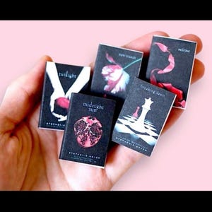 Puede incluir: Conjunto de libros en miniatura de la saga Crepúsculo de Stephenie Meyer. Las portadas muestran imágenes icónicas de los libros, incluyendo una granada, una pieza de ajedrez y diseños florales, sobre un fondo oscuro. Los libros están en una mano.
