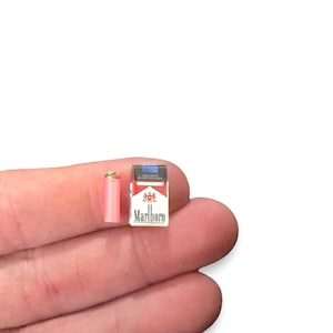 Pode incluir: Um maço de cigarros em miniatura vermelho e branco com o texto Marlboro impresso na frente, acompanhado por um minúsculo isqueiro cilíndrico rosa com topo metálico, ambos em escala sobre a ponta de um dedo.