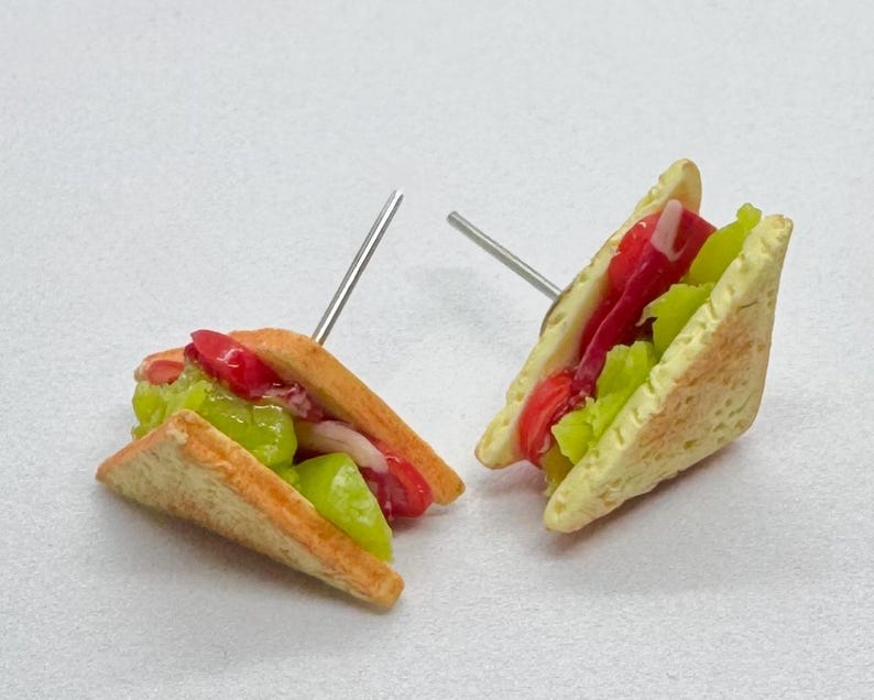 Pu&ograve; includere: Un paio di orecchini a forma di sandwich in miniatura. Ogni orecchino presenta un sandwich triangolare con strati di pomodori rossi, lattuga verde e altri ripieni tra pane tostato, tenuti insieme da un perno d'argento.
