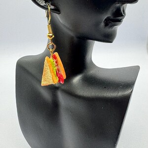 Pu&ograve; includere: Orecchini pendenti color oro con ciondoli a forma di sandwich in miniatura. Ogni ciondolo &egrave; un sandwich triangolare con strati di pane, lattuga, pomodoro e altri ripieni. Gli orecchini sono attaccati a una testa di manichino nera.