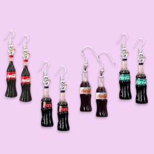 以下が含まれることがあります： ダークブラウンのガラス製ミニチュアボトルチャームが付いた4組のドロップピアス。各ボトルには、赤いコカ・コーラのロゴが入った白いラベルが貼られています。チャームは、小さな装飾ループが付いた銀色の金属製フィッシュフックピアス金具に取り付けられています。