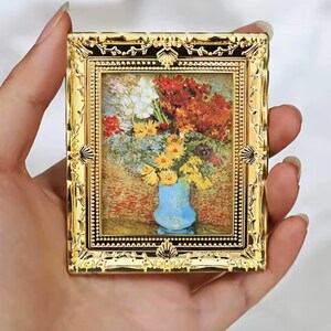 May include: A miniature rectangular picture frame with a shiny gold finish and ornate, embossed detailing. The frame encloses a small, detailed print of a vibrant floral bouquet in a light blue vase, featuring red, yellow, and white flowers.