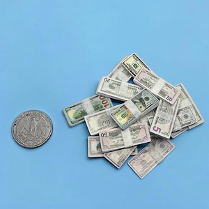 May include: A collection of miniature toy US dollar bills bundled with white paper bands, featuring various denominations including 5, 20, 50, and 100 dollar notes, positioned next to a single silver-coloured US quarter coin for scale comparison.