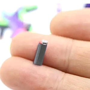 May include: A miniature cylindrical object with a dark gray body and a small silver-colored top cap. The item is held between two fingers, showcasing its tiny scale and simple, smooth, vertical design with a subtle red accent near the top.