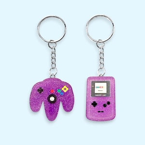 May include: Two purple glitter keyrings. One is a controller with colourful buttons, the other is a handheld game console. Both have silver key rings and chains. The game console keyring has the word "GAME B" on the screen.