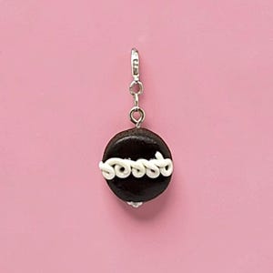 May include: A small, round, dark brown charm shaped like a cookie or cake, featuring a horizontal line of white, swirled icing across the centre. The charm is attached to a silver-toned metal lobster clasp by a small circular jump ring.
