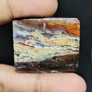 May include: A square-cut gemstone with a unique, layered pattern. The stone features bands of color, including orange, brown, yellow, and white, creating a landscape-like appearance. The gemstone is held between two fingers.