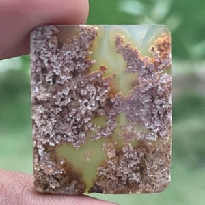 May include: A rectangular gemstone with a light green and brown pattern. The stone has a textured, crystallized appearance with areas of light green and brown. The edges are slightly rounded, and the stone is held by a finger.