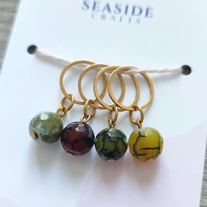 Peut inclure: Quatre marqueurs de point dorés avec des perles colorées à motifs : vert, bordeaux, vert à motifs et jaune. Les marqueurs sont attachés à une carte blanche avec le texte "SEASIDE CRAFTS".