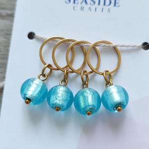 Könnte beinhalten: Vier goldfarbene Maschenmarkierer mit hellblauen, gläsernen Perlen. Die Perlen haben ein schimmerndes, strukturiertes Aussehen. Die Marker sind an einer weißen Karte mit dem Text "SEASIDE CRAFTS" und einem Fischlogo befestigt.