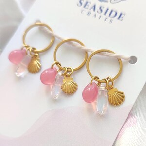 Op de afbeelding: Drie goudkleurige metalen ringen bevestigd op een witte displaykaart met de tekst SEASIDE CRAFTS en een blauw vislogo. Elke ring heeft drie bungelende bedels: een roze kraal, een doorzichtige druppelvormige kraal en een kleine goudkleurige schelp.