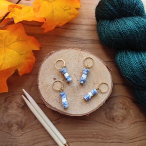 Op de afbeelding: Vier steekmarkeerders, elk bestaande uit een goudkleurige metalen ring bevestigd aan een verticale reeks kleine, platte, cilindrische kralen. De kralen hebben een herhalend patroon van gemarmerd blauw en wit voor een duidelijk kleurcontrast.