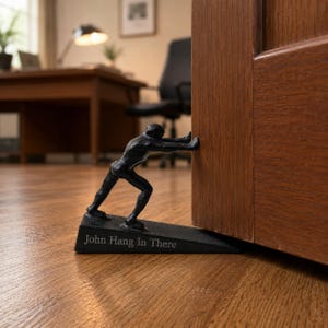Pode incluir: Um batente de porta de ferro fundido preto em forma de figura humana empurrando uma porta de madeira. A base em cunha apresenta o texto gravado "John Hang In There" numa fonte clara, servindo como um acessório funcional e decorativo.