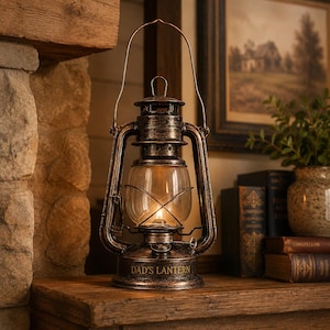 Peut inclure: Une lanterne tempête en métal aux tons bronze avec un globe en verre transparent et une poignée en fil de fer. La base porte l'inscription gravée DAD'S LANTERN. La lanterne contient une petite source lumineuse visible à travers le verre.