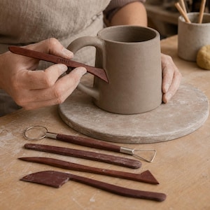 May include: A grey clay mug being smoothed with a dark brown wooden tool engraved with the name EMMA. Below the mug are four additional pottery tools: a wire loop tool, a flat wooden stick, a pointed wooden tool, and a wooden rib tool.