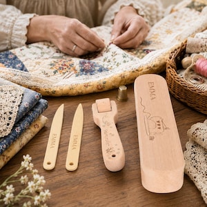 May include: A set of light-colored wooden sewing tools including two point turners, a seam roller, and a rectangular storage box. Each item is engraved with the name EMMA. The seam roller features floral designs and the text Seam roller.