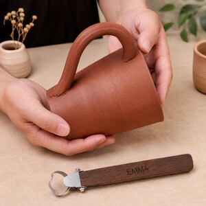 Puede incluir: Una taza de arcilla color terracota con un asa grande y curva y una herramienta de alfarería de madera con una hoja de bucle de metal y el nombre EMMA grabado en el mango de madera marrón oscuro, sobre una superficie de tela clara.