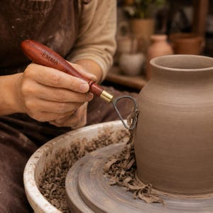 May include: A pottery trimming tool with a dark reddish-brown wooden handle engraved with the name EMMA and a brass ferrule connecting to a teardrop-shaped metal loop blade, shown carving a grey clay vessel on a spinning pottery wheel.