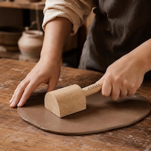 May include: A light-colored wooden pottery mallet with a semi-cylindrical head and a handle engraved with the name EMMA, positioned on a flat, circular piece of brown clay. The tool features a smooth, natural wood grain finish throughout.