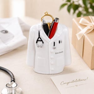 Peut inclure: Un organisateur de bureau en céramique blanche en forme de blouse de laboratoire avec un motif de stéthoscope noir, un anneau doré et le nom Ken Smith imprimé sur le devant. Il contient un stylo noir, un stylo doré et un stylo rouge.
