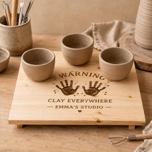 May include: A light-coloured wooden board featuring three small, empty clay bowls. The board is engraved with the text "WARNING CLAY EVERYWHERE EMMA'S STUDIO" and two handprints. The surface has a natural wood grain and a smooth, flat finish.