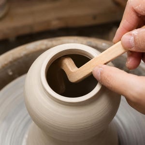 Può includere: Uno strumento da vasaio in legno chiaro con un profilo curvo a forma di S, utilizzato per modellare il collo interno di un vaso di argilla umida grigio chiaro su un tornio, evidenziando la consistenza liscia del legno e dell'argilla.