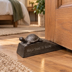 May include: A dark brown, wedge-shaped doorstop made of cast metal featuring a sculpted turtle on top. The front face of the wedge is engraved with the text "MILO TAKE IT SLOW" in capital letters. The object is positioned to hold a wooden door open.
