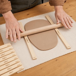 Könnte beinhalten: Ein hellbraunes Nudelholz aus Holz glättet ein Stück beigefarbenen Ton, das zwischen zwei Holzleisten mit der Aufschrift EMILY und 7mm liegt. Ein Stapel weiterer Holzleisten mit Dickenangaben von 3mm bis 12mm befindet sich daneben.