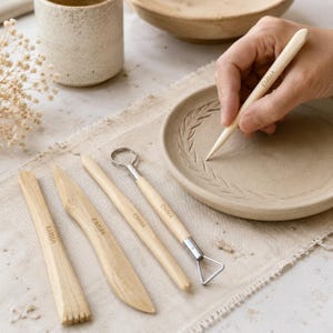 May include: A set of light-colored wooden pottery tools, each engraved with the name EMMA. The collection includes a flat modeling tool, a knife, a pointed stylus, and a wire loop tool, all used for shaping and detailing a circular clay plate.