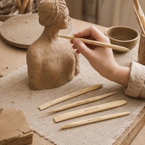 Könnte beinhalten: Ein Set aus fünf hellen Töpferwerkzeugen aus Holz, die jeweils mit dem Schriftzug EMMA POTTERY versehen sind. Die Werkzeuge haben verschiedene spitze, flache und abgerundete Enden zum Formen von Ton, neben einer Tonbüste.