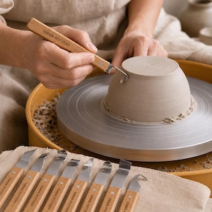 Może przedstawiać: Zestaw narzędzi garncarskich z jasnymi drewnianymi uchwytami, każdy oznaczony napisem EMMA POTTERY. Jedno narzędzie jest używane do wycinania cienkiego paska gliny z podstawy odwróconej, jasnoszarej misy glinianej umieszczonej na metalowym kole.