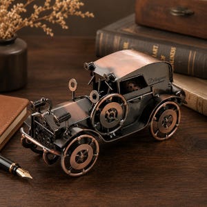 May include: A vintage-style metal car model crafted from dark and copper-toned components, featuring circular wheels, a prominent front grille, and the word "Děkujeme" engraved on the side panel. The structure is assembled with visible screws.