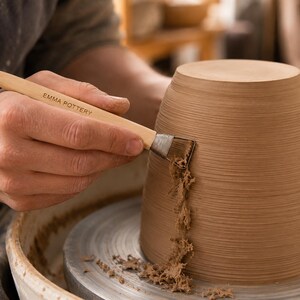 May include: A light-colored wooden pottery tool with the text EMMA POTTERY printed on the handle, featuring a metal ferrule and a serrated blade edge, being used to trim excess clay from the side of a brown, ribbed, inverted ceramic vessel.