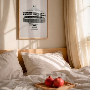 May include: A framed architectural print in a bedroom setting. The artwork, a black and white illustration of a building, is displayed above a bed with white linens. Two pomegranates rest on a wooden tray. The frame is made of oak.